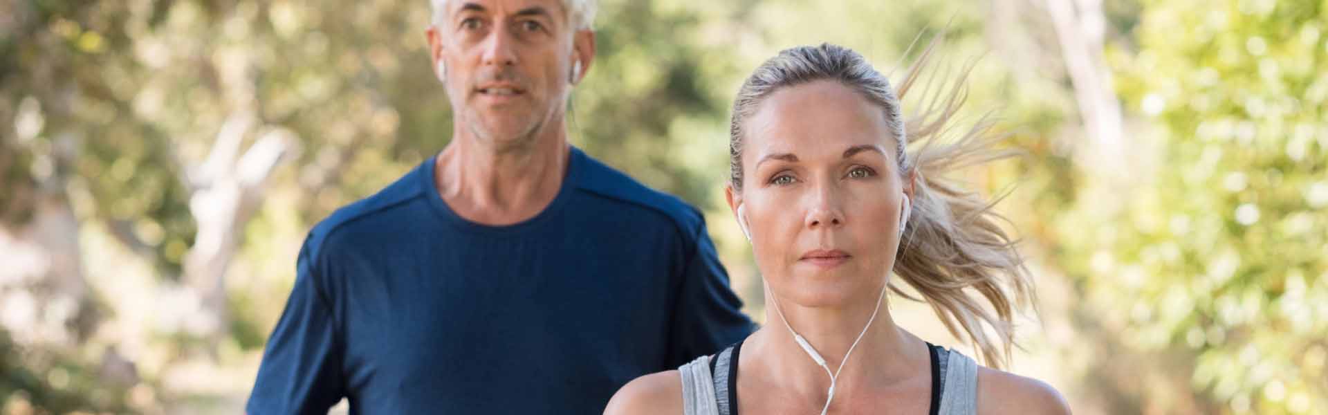 A man and woman jogging together in a park, surrounded by greenery and a clear blue sky.